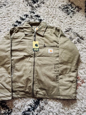 Beige overshirt/jacka från Carhartt - Snygg beige overshirt från Carhartt med dragkedja framtill, klassisk krage och två sidofickor. Bröstficka med Carhartt-logga och innerfoder i quiltad polyester. Materialet är slitstark bomull och polyester, perfekt för en streetwear-look.