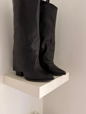 Svarta höga boots med klack - Snygga svarta stövlar i slätt konstläder med hög, bred klack och spetsig tå. Stövlarna har en stilren design och går upp till knäna. Perfekta för att lyfta vilken outfit som helst. Säljer då de inte kommer till användning, lite slitna men syns inte när man bär dem. Perfekta till kjol!