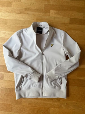 Vit bomberjacka från Lyle & Scott - Snygg vit bomberjacka från Lyle & Scott med klassisk gul logga på bröstet. Jackan har dragkedja framtill, två fickor med dragkedja och ribbade muddar vid ärmar och midja. Perfekt för dig som gillar stilren och sportig look.