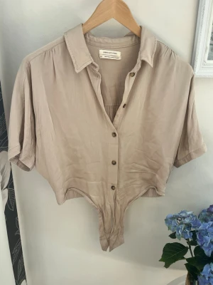 Beige kortärmad blus från Urban Outfitters - Stilren beige knytblus från Urban Outfitters med korta ärmar och klassisk skjortkrage. Modellen har knappar framtill och är tillverkad i ett mjukt, tunt tyg som ger en avslappnad känsla. Perfekt att matcha med jeans eller kjol för en trendig look.