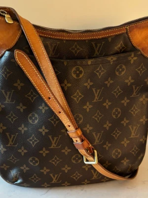 Louis Vuitton Monogram axelväska - Louis Vuitton Odeon MM axelväska i brunt monogram-canvas.  Tillverkad Frankrike år 2008. Storlek ca 30x30 cm.   Väskan har justerbar axelrem och detaljer i vachetta-läder.Den har en dragkedja som stängning och en öppen ficka på framsidan. Väl använd men hel.   