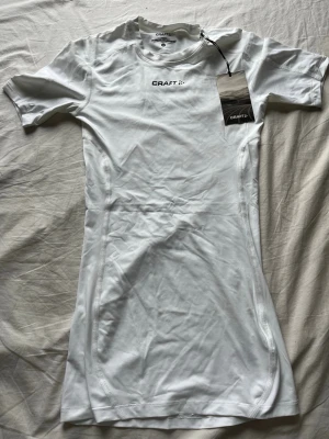 Vit compression shirt från Craft - Snygg vit träningströja/compression shirt från Craft med korta ärmar och rund hals. Tillverkad i ett stretchigt och fukttransporterande material som passar perfekt för intensiva träningspass. Diskret logga på bröstet och liten detalj på ryggen. Den är också helt oanvänd.