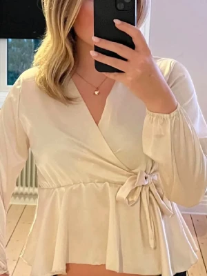 Beige omlottblus med knytband - Snygg beige omlottblus med knytband i midjan och v-ringning. Blusen har långa ärmar med resår vid ärmslut och en lätt volang nertill. Perfekt för en trendig och stilren look.