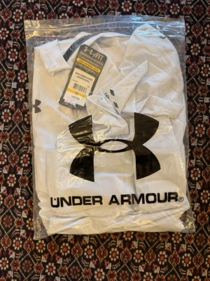Vit träningströja från Under Armour - Snygg vit t-shirt från Under Armour med svart logga på bröstet och tryckt varumärke längs sidan. Tillverkad i ett lätt och snabbtorkande funktionsmaterial som passar perfekt till träning och sportaktiviteter. Storlek M passar S. OBS defekt på bild nr 3 lite fläckig på ärmen och lite där bak därför jag vill sälja den!
