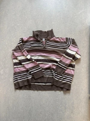 Randig stickad tröja med dragkedja - Mysig stickad tröja med bruna, rosa och vita ränder. Tröjan har halv dragkedja vid halsen och ribbstickad krage samt muddar. Snygg och avslappnad passform. Tunnare material så den passar perfekt nu mot sommarkvällar. Sitter som M. 