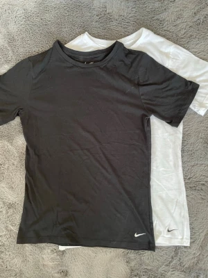 Svart och vit Nike t-shirt duo - Två klassiska t-shirts från Nike, en svart och en vit, båda med liten Nike-logga nere vid fållen. T-shirtsen har rund hals och korta ärmar, tillverkade i mjukt material som känns skönt mot huden.