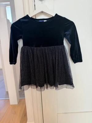 Svart sammetsklänning med glittertyll - En svart klänning med sammetsliv och långärmade ärmar. Kjolen är i tyll med glittriga prickar som ger en festlig känsla. Perfekt för dig som vill sticka ut lite extra. Rund halsringning och normal passform.