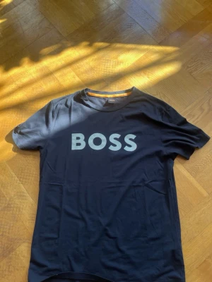 Svart BOSS t-shirt med tryck - Svart t-shirt från BOSS med stort guld BOSS-tryck på bröstet. Klassisk rund halsringning och korta ärmar. Tillverkad i mjuk bomull som känns skön mot huden. Perfekt för en clean och stilren look.