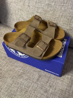 Birkenstock tofflor  - Klassiska Birkenstock Arizona sandaler i brun mocka med två breda remmar och justerbara metallspännen. Fotbädden är ergonomiskt utformad för extra komfort och sulan är i kork. Perfekta för en avslappnad och trendig stil.