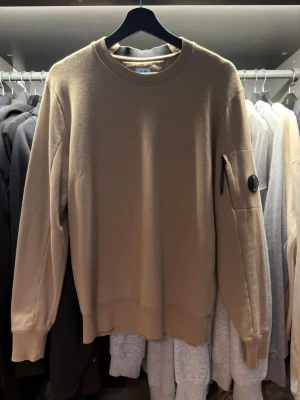 Beige sweatshirt från C.P. Company - Stilren beige sweatshirt från C.P. Company med rund halsringning och ribbade muddar. Tröjan har en ikonisk linsdetalj på ärmen och en praktisk ficka med dragkedja. Storlek S, pris kan diskuteras