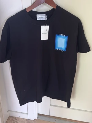 Svart Casablanca t-shirt med blå tryck - Svart t-shirt från Casablanca med grafiskt blått tryck och texten 'Casa Blanca' både fram och bak. Klassisk rund hals och korta ärmar. Perfekt för dig som gillar streetwear och vill sticka ut med en snygg design.