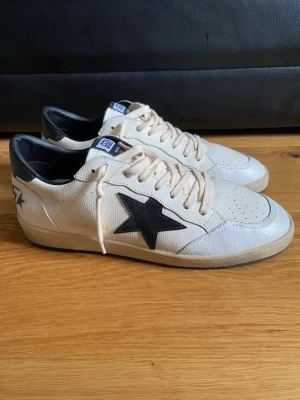 Golden Goose Ballstars vita sneakers med svart stjärna - Golden Goose sneakers i vitt läder med svart stjärna på sidan och svarta detaljer vid hälen. Snygg text 'SNEAKERS' på baksidan och klassisk låg modell. Perforerad tå och beige sula ger en cool vintagekänsla. Perfekta för dig som gillar streetstyle.