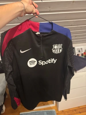 Svart FC Barcelona träningsjacka Nike - Säljer en svart träningsjacka från Nike med FC Barcelona-emblem och Spotify-logga på bröstet. Jackan har dragkedja framtill, röda och blå detaljer på axlarna och är tillverkad i ett lätt syntetmaterial. Perfekt för fotbollsträning eller sport.