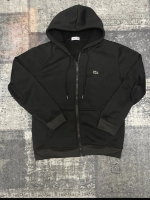 Svart hoodie från Lacoste med dragkedja - Snygg svart hoodie från Lacoste med dragkedja framtill och klassisk krokodillogo på bröstet. Regular fit med huva och snörning, samt ribbade muddar vid ärmar och nederkant. Perfekt för en chill och stilren look.