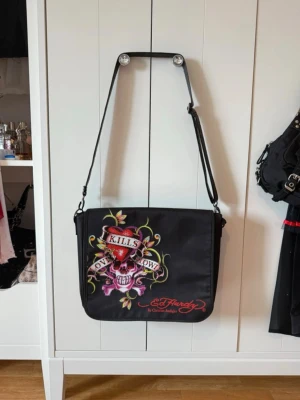 Svart Ed Hardy axelväska😱🙏🙏 - Säljer en svart axelväska från Ed Hardy med stort färgglatt tryck av hjärta, dödskalle och texten 'Love Kills Slowly' på framsidan. Väskan har justerbar axelrem och stängs med kardborre. Insidan är fodrad med rött Ed Hardy-tryck. Säljer för kommer inte till användning har bara använts ett fåtal gånger.😛🙏❤️❤️❤️