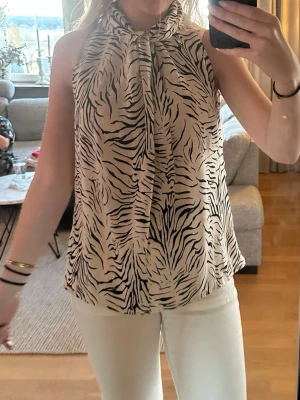 Zebramönstrad ärmlös blus med knyt - Snygg ärmlös blus med zebramönster i svart och vitt. Toppen har hög hals med knyt och en liten öppning i ryggen. Luftig och lätt i materialet, perfekt för en trendig look.