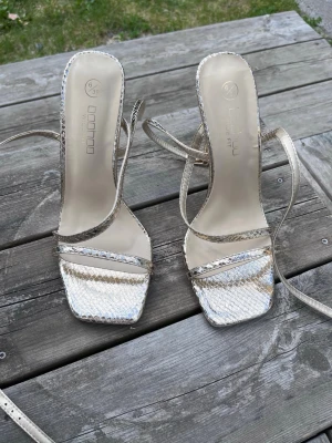 Guldiga pumps med snörning från Bohoo - Helt oanvända kackar med plasten kvar på. Säljer ett par trendiga guldiga pumps från bohoo med smala remmar och snörning runt vristen. Skorna har fyrkantig tå, hög klack och en snygg ormskinnsmönstrad yta som ger extra edge till outfiten.
