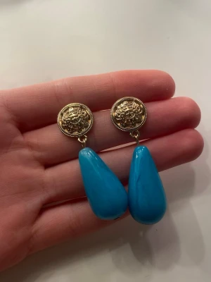 Blå droppformade örhängen med gulddetalj - Snygga örhängen med rund guldfärgad knopp med mönster och stora, blanka droppformade hängen i klarblå plast. Perfekta för att ge din outfit en färgklick och cool retrovibe. Stiftmodell för enkel användning. Helt oanvända ❤️
