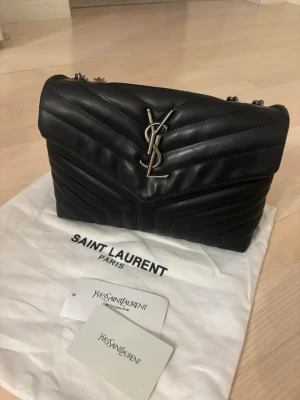 Svart väska från YSL - Snygg svart axelväska från Saint Laurent med quiltat mönster och silverfärgad YSL-logga framtill. Väskan har kedjeremmar i metall och är tillverkad i mjukt läder. Perfekt för dig som vill ha en lyxig och trendig accessoar.