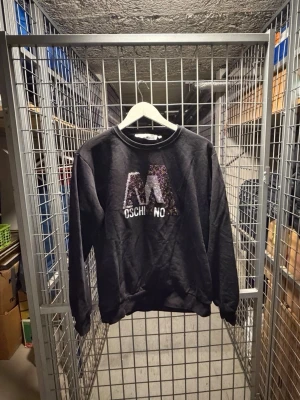 Svart Moschino sweatshirt med paljetter - Svart sweatshirt från Love Moschino med stort M i paljetter och text på bröstet. Rund halsringning och ribbade muddar. Söt nallepatch på ena ärmen. Perfekt för dig som gillar streetstyle och vill sticka ut lite extra.