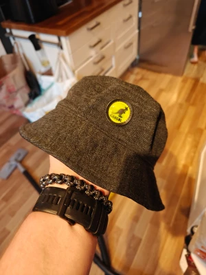 Grå bucket hat från Kangol - Säljer en grå bucket hat från Kangol med en rund gul och svart logga framtill. Hatten är i mjukt stickat material och har klassisk form med nedvikt brätte. Perfekt accessoar för en avslappnad streetstil.