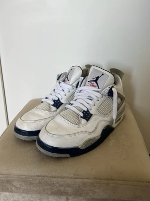 Air Jordan 4 Retro Vit/Marinblå - Snygga Air Jordan 4 Retro sneakers i vitt läder med marinblå och grå detaljer. Klassisk siluett med meshpaneler, Jumpman-logga på plösen och hälen samt vita skosnören. Perfekta för dig som gillar streetwear och ikoniska sneakers.