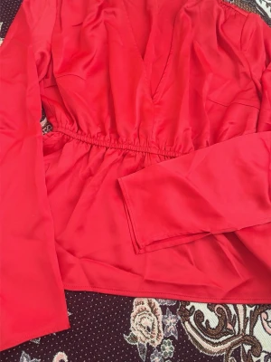 Röd omlottblus med lång ärm - Trendig röd omlottblus med markerad midja och långa ärmar. Blusen har en djup v-ringning och är tillverkad i ett mjukt, glansigt tyg som ger en snygg look. Perfekt för att lyfta vilken outfit som helst.