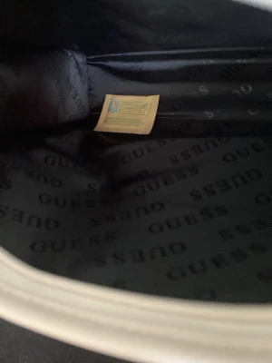 Ljusrosa axelremsväska från Guess - Helt ny Snygg ljusrosa handväska från Guess med vit logotypprint över hela väskan och vita detaljer. Väskan har en klassisk rektangulär form, justerbar axelrem och Guess metallogo framtill. Materialet är konstläder som ger en lyxig känsla och trendig look.