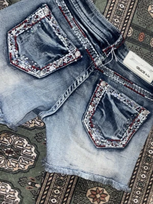 Blå jeansshorts med broderade fickor - Unika blå jeansshorts från Chillaz med broderade detaljer och pärlor på bakfickorna. Shortsens kanter är råa och fransiga för en avslappnad look. Röd och vit brodyr längs fickor och midja ger extra edge. Perfekta för sommardagar och festivaler.