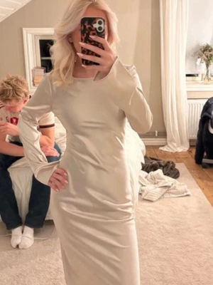 Lång ljus sidenklänning med rosett - Elegant långklänning i ljus sidenliknande material med lång ärm, öppen rygg och stor rosettdetalj bak. Klänningen har en smickrande passform och slät yta som ger ett lyxigt intryck. Perfekt för dig som vill sticka ut med en stilren look.