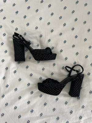 Svarta platåsandaler med vita prickar - Säljer ett par helt oanvända ny med lappen kvar svarta sammet platåsandaler från M&S med vita prickar och bred 13cm högt klack. Sandalerna har en söt rosett framtill, justerbart ankelband med spänne och är tillverkade i ett sammetsliknande material. Perfekta för dig som vill sticka ut lite extra.