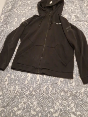 Svart hoodie från Sail Racing - Svart hoodie från Sail Racing med dragkedja framtill och två stora fickor. Har tryckt logga på bröstet och ärmen samt justerbar huva med snören. Tillverkad i mjukt bomullsmaterial, perfekt för en avslappnad och sportig stil. Vänstra ärmen har en super liten skada som inte syns men annars nyskick.