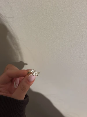 Guldfärgad ring med hjärnor  - Säljer en guldfärgad ring med två hjärter som korsar varandra med dekoration framtill. Ringen har gnistrande stenar både på blomman och längs bandet, vilket ger en lyxig och trendig känsla. Perfekt för dig som gillar unika och glittriga accessoarer.