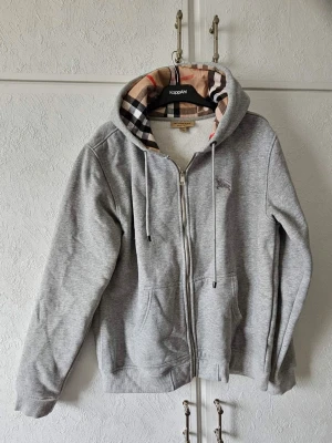 Grå zip hoodie med Burberry-mönster - Snygg grå hoodie med dragkedja från Burberry. Klassisk rutig insida i huvan och två fickor framtill. Mjuk bomullsblandning och justerbar huva med snören. Perfekt för en avslappnad och trendig look. (Står 2XL men sitter igentligen som en M/L, OBS r3pl1ka)