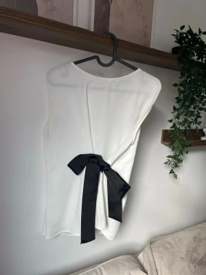 Vit ärmlös topp med svart rosett - Stilren vit ärmlös topp med rund halsringning och en snygg svart rosett i midjan baktill. Toppen har en enkel, rak passform och är tillverkad i ett mjukt, lätt material som ger en clean look. Perfekt för dig som gillar minimalistisk stil med en twist.