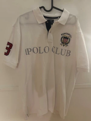 Vit Polo Club pike Kingshill - Vit pikétröja från Kingshill Polo Club med klassisk pikékrage och knappslå. Stor brodyr med logga och text på bröstet samt en stor röd siffra 3 på ärmen. Perfekt för en sportig och stilren look.