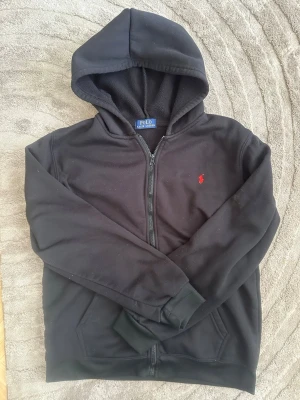 Svart hoodie från Polo Ralph Lauren - Svart hoodie med dragkedja från Polo Ralph Lauren. Klassisk design med huva, två fickor framtill och det ikoniska röda Polo-märket broderat på bröstet. Tillverkad i mjukt och bekvämt material, perfekt för en avslappnad stil.