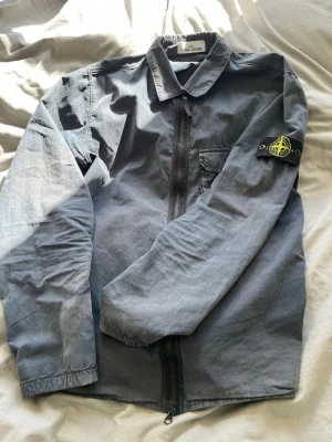  Äkta Marin blå overshirt från Stone Island - Snygg ÄKTA marin blå overshirt från Stone Island med klassisk krage och dragkedja framtill. Har en bröstficka och den ikoniska Stone Island-patchen på vänster ärm. Tillverkad i ett lätt bomullsmaterial som ger en avslappnad men stilren look. Finns dock lite tecken på slitage och ett lagad hål vid insidan av höger armen men det syns ej när man har på. Priset kan diksuteras!! Och skriv vid minsta lilla frãga
