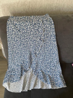 Blommig blå omlottkjol med volang - Söt blå långkjol med vitt blommönster och omlottdesign. Kjolen har volangkant nedtill som ger en extra touch och knytband i midjan. Perfekt för dig som gillar en feminin och somrig stil.