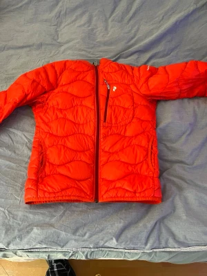 Röd dunjacka från Peak Performance - Säljer en riktigt snygg röd dunjacka från Peak Performance. Jackan har en cool quiltad design, dragkedja framtill och fickor med dragkedja. Perfekt för kalla vinterdagar och håller dig varm med sitt lätta dunmaterial.