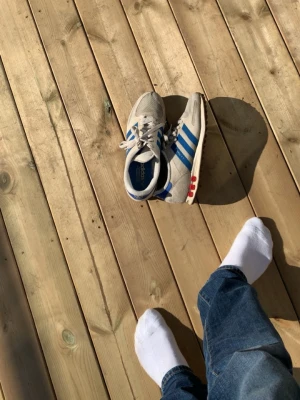 Adidas beige sneakers med blå ränder - Säljer ett par klassiska Adidas sneakers i beige med blå ränder på sidorna och röda detaljer vid hälen. Skorna har snörning, låg profil och en sula med bra grepp. Perfekta för dig som gillar retrostil och sportig look.