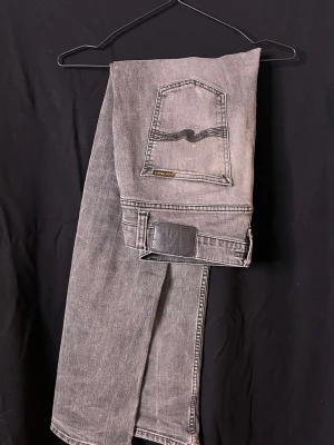 Grå jeans från Nudie Jeans - Säljer ett par gråa jeans från Nudie Jeans med klassisk femficksmodell och svarta sömmar på bakfickan. Jeansen har en rak passform och är tillverkade i slitstark denim. Perfekta för dig som gillar en avslappnad men ändå stilren look.