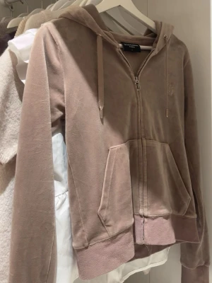  Juicy Couture - Mjuk beige hoodie i velour från Juicy Couture med dragkedja, huva och fickor framtill. Klassisk logga broderad på bröstet och ribbade muddar vid ärmslut och nederkant. Perfekt för en chill och trendig look.