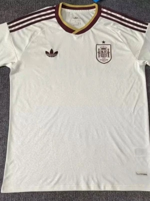 Vit Spanien Adidas fotbollströja - Snygg vit fotbollströja från Adidas med Spaniens landslagsemblem på bröstet. Tröjan har vinröda detaljer, klassiska Adidas-ränder på axlarna och gul/vinröd krage. Tillverkad i lätt och ventilerande material med Climalite-teknologi. Du kan bestämma vilken storlek du vill ha!!! Det kan ta upp till 15-20 dagar för tröjorna att komma!!! 