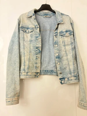 Ljusblå klassisk jeansjacka - Säljer en ljusblå jeansjacka i klassisk modell med två  med lock och knapp, samt silverfärgade knappar framtill. Jackan har en rak passform och är tillverkad i denim med tvättad effekt för en cool vintagekänsla. Perfekt att slänga över en hoodie eller t-shirt.