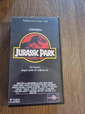 Jurassic Park VHS-film - Klassisk VHS-utgåva av filmen Jurassic Park, regisserad av Steven Spielberg. Svart plastfodral med ikoniskt rött och gult dinosaurielogo på framsidan. Text och bilder på baksidan, svensk text och tal. Perfekt för samlare av retrofilm eller filmnostalgi.