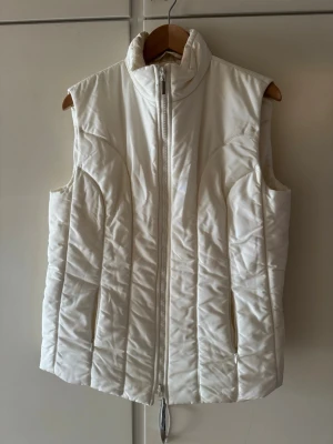 lusia cerano Vit dunväst  - Stilren vit dunväst med hög krage och hel dragkedja framtill. Västen har en quiltad design med vertikala sömmar och är ärmlös för en clean look. Perfekt att slänga över en hoodie eller tröja när det är kyligt ute. Materialet är glansigt och ger en fräsch vibe.