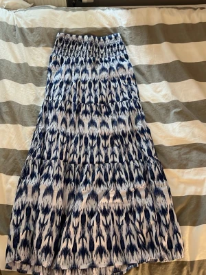 Blåvit långkjol med mönster - Säljer en blåvit långkjol med elastisk midja och ett snyggt abstrakt mönster. Kjolen har en luftig passform och är perfekt för varmare dagar. Materialet känns lätt och bekvämt mot huden.
