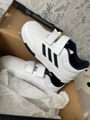 Vita Adidas sneakers med kardborre - Snygga vita sneakers från Adidas med klassiska svarta ränder på sidan och två kardborreband för enkel påtagning. Skorna har svart innerfoder och perforerade detaljer på sidan för extra ventilation. Perfekta för en sportig och clean look. Oanvända