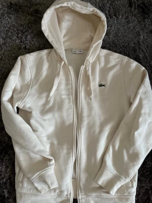 Vit hoodie med dragkedja från Lacoste - Snygg vit hoodie från Lacoste med dragkedja framtill och klassisk krokodil-logga på bröstet. Hoodien har justerbar huva med snören och ribbade muddar vid ärmar och nederkant. Tillverkad i mjukt bomullsmaterial för skön känsla.
